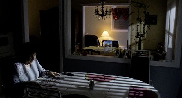 Still from the film "Happer’s Comet" by Tyler Taormina. An elderly woman sits at a table in the dark apartment. The light shining in from outside casts a striped pattern on the table.