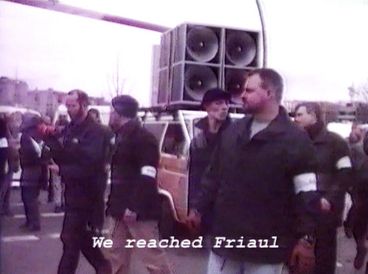 Filmstill aus „Normalität 1–10“ von Hito Steyerl. Menschen auf einer Demo, ein Lautsprecherwagen und Ordner. 