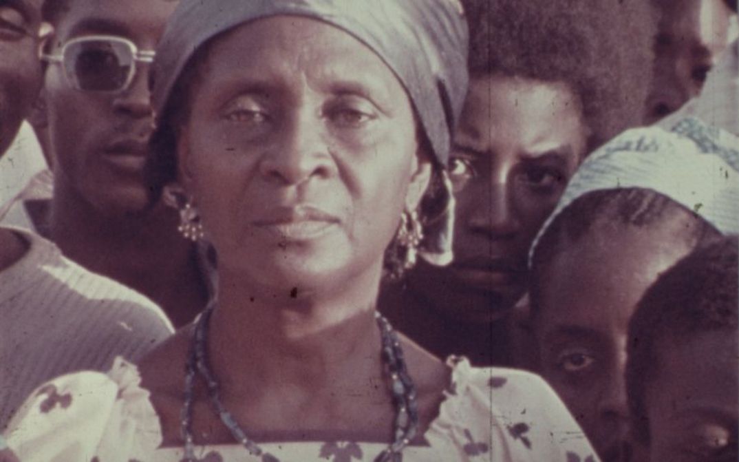 Filmstill aus The Return of Amílcar Cabral: Eine Frau blickt mit ernstem Gesicht in die Kamera. Um sie herum stehen weitere Menschen.
