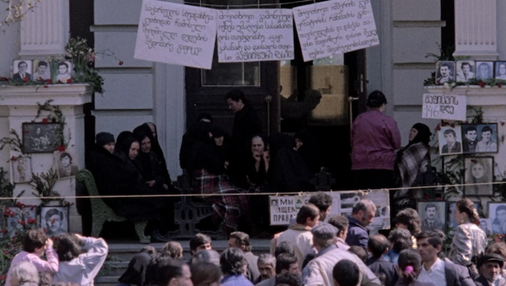 Filmstill aus The Way Home – Merab Mamardashvili: Menschen bei einer Kundgebung in Tbilisi. Man sieht neben den Menschen aufgehängte Plakate in georgischer Schrift und Fotos von Menschen.