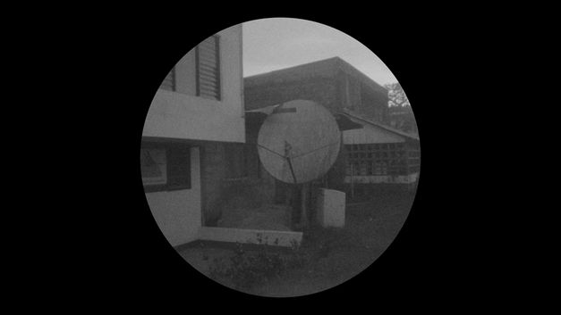 Filmstill aus dem Film "Yarokamena" von Andrés Jurado. Rundes Schwarz-Weiß-Bild einer Satellitenschüssel, im Hintergrund stehen Häuser.