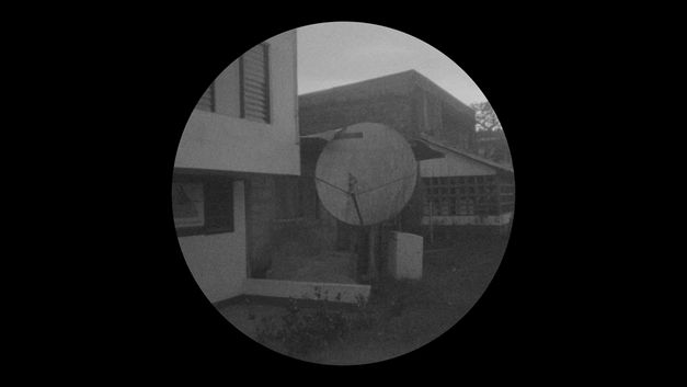 Still from the film "Yarokamena" by Andrés Jurado. Round black and white image of a satellite dish next to houses.