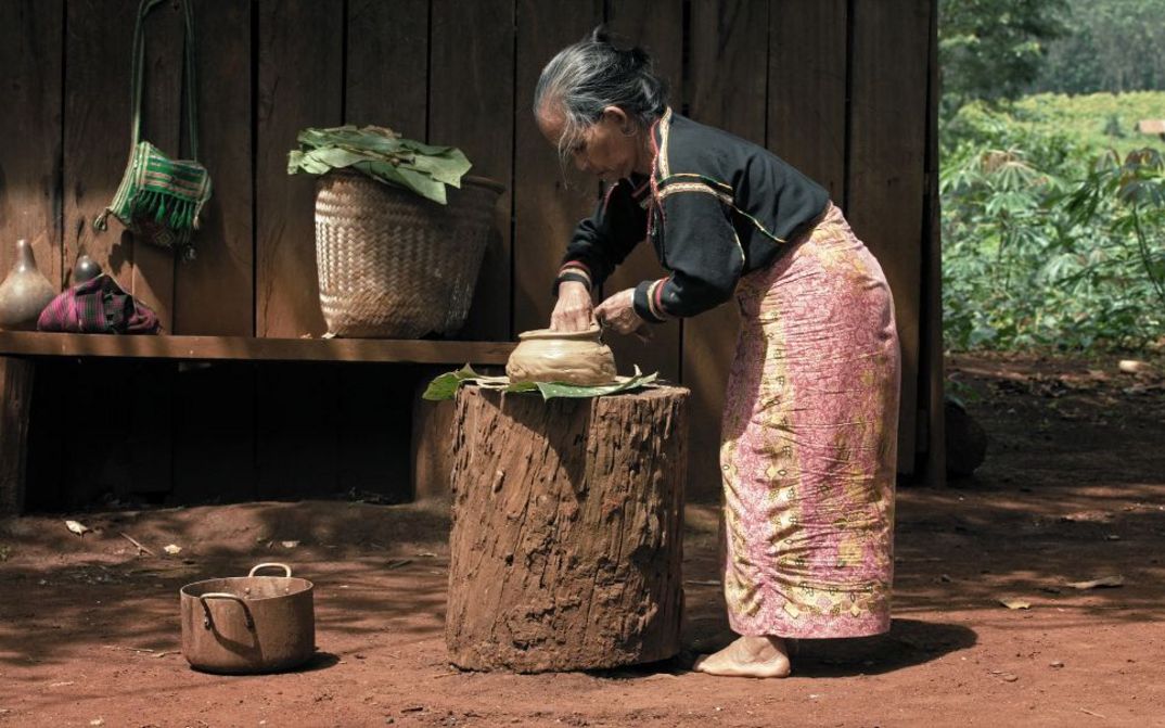 Ältere Frau bereitet auf einem Holzblock Teig oder Speisen zu, umgeben von Körben und Gefäßen vor einer Holzwand im Freien.