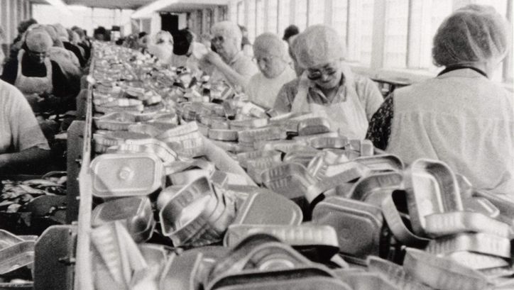 Filmstill aus BELFAST, MAINE. Frauen arbeiten am Fließband einer Fabrik, in der Mitte sieht man einen Haufen leerer Konservendosen.