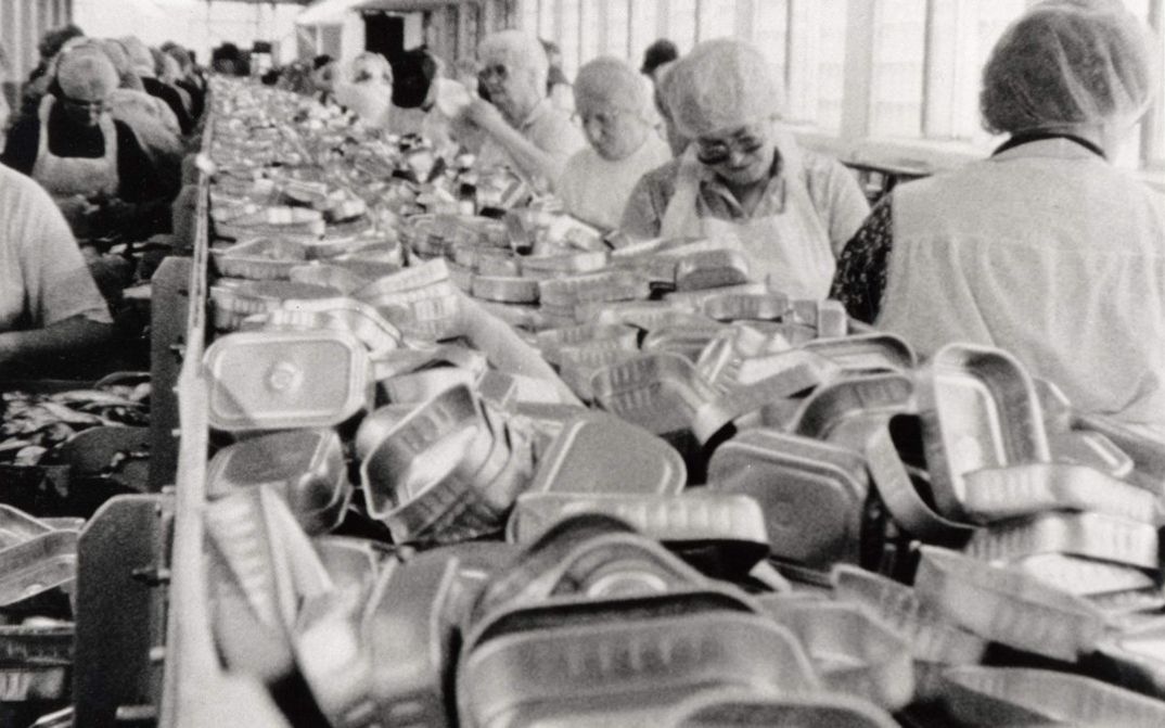 Filmstill aus BELFAST, MAINE. Frauen arbeiten am Fließband einer Fabrik, in der Mitte sieht man einen Haufen leerer Konservendosen.