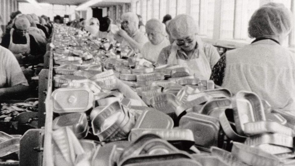 Filmstill aus BELFAST, MAINE. Frauen arbeiten am Fließband einer Fabrik, in der Mitte sieht man einen Haufen leerer Konservendosen.