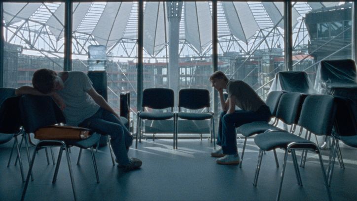 Film still from CENTER: A few chairs in a room at the DFFB, in the background you can see the roof of the Sony Center through the glass pane. Two men sit bored on the chairs.