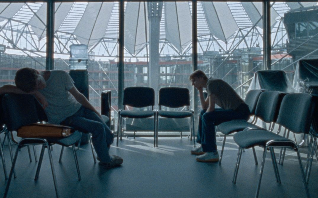 Film still from CENTER: A few chairs in a room at the DFFB, in the background you can see the roof of the Sony Center through the glass pane. Two men sit bored on the chairs.