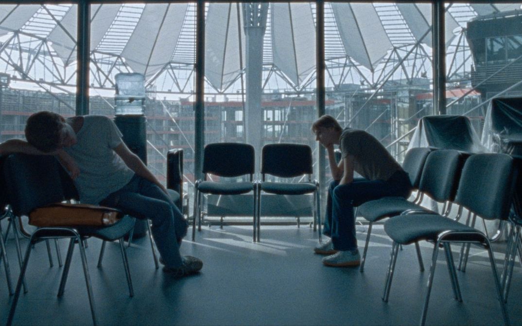 Film still from CENTER: A few chairs in a room at the DFFB, in the background you can see the roof of the Sony Center through the glass pane. Two men sit bored on the chairs.