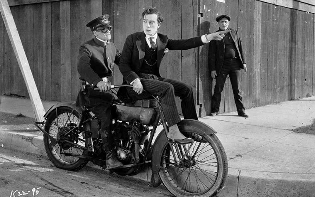 Film still from SHERLOCK JUNIOR: Buster Keaton, wearing an elegant suit, is sitting on the handlebars of a motorcycle and pointing the way with one hand. Behind him sits the driver in uniform.