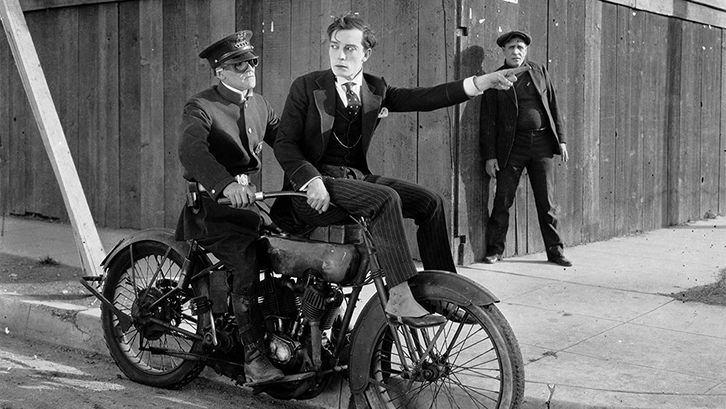 Film still from SHERLOCK JUNIOR: Buster Keaton, wearing an elegant suit, is sitting on the handlebars of a motorcycle and pointing the way with one hand. Behind him sits the driver in uniform.