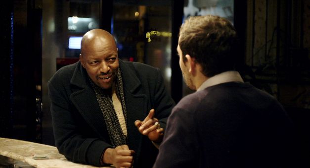 Still from the film "Une fleur à la bouche" Éric Baudelaire. A bald man in a dark green overcoat leaning on a bar points coyly to a man he is speaking with. 