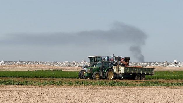 Traktor mit Anhänger auf grünem Feld, Rauch steigt über Stadt im Hintergrund.