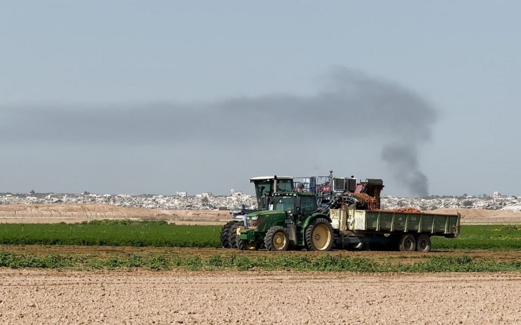 Traktor mit Anhänger auf grünem Feld, Rauch steigt über Stadt im Hintergrund.