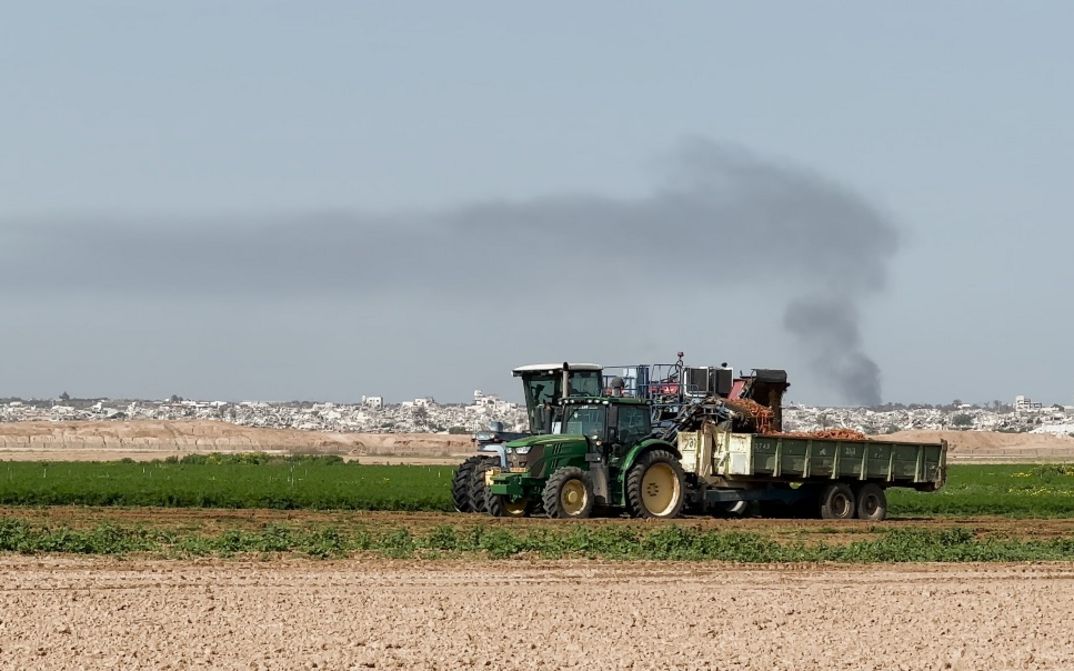 Traktor mit Anhänger auf grünem Feld, Rauch steigt über Stadt im Hintergrund.