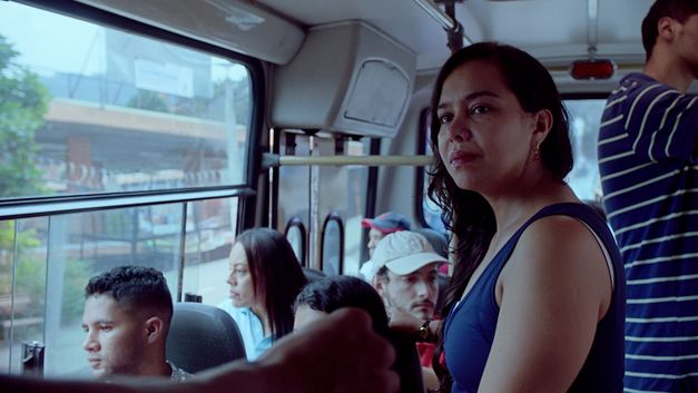 Film still from "La piel en primavera" by Yennifer Uribe Alzate. It shows a close-up of a standing woman in a bus. The bus is filled with people. 