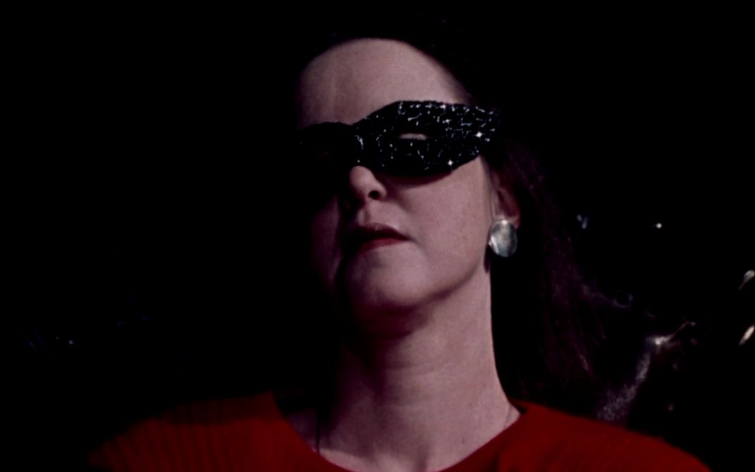 Film still from CHANGE OF FATE: You see the face of a woman with a black mask around her eyes.