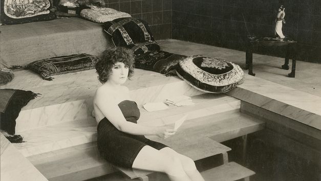 Filmstill aus LA BELLE DAME SANS MERCI: Eine junge Frau sitzt auf der Treppe in einem glamourösen Haus.
