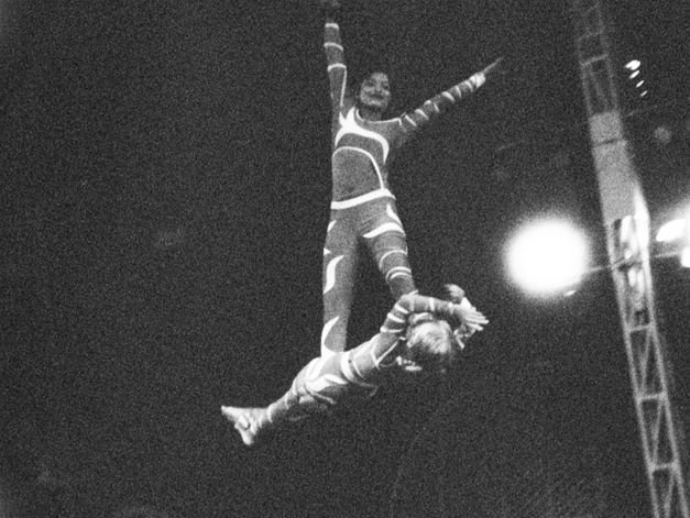 Film still from Gavati Wad’s film “O Seeker”. Two trapese artists wear stripey costumes as they perform. One of them supports the other, who balances on top of them and smiles, both arms up in the air. 