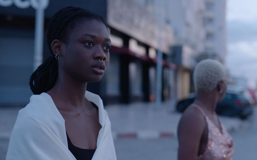 Filmstill aus PROMIS LE CIEL: Zwei junge Frauen stehen an einer Straße, der Hintergrund ist verschwommen.