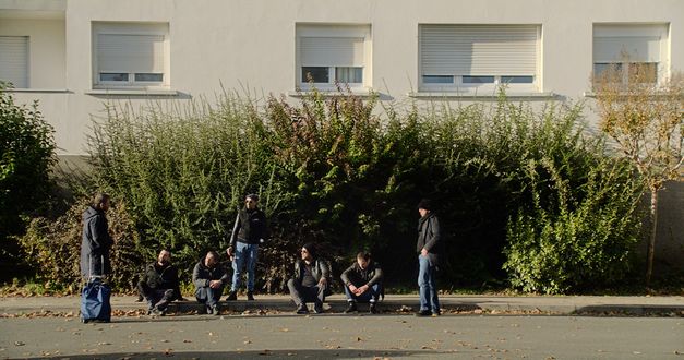 Filmstill aus dem Film "Le Gang des Bois du Temple" von Rabah Ameur-Zaïmeche. Vier Männer in dunkler Kleidung sitzen auf einem Bordstein, drei Personen stehen um sie herum und schauen sich gegenseitig an. Im Hintergrund steht ein grüner Busch vor einem Haus mit Fenstern und halb verschlossenen Rollos.