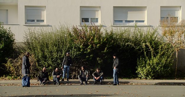 Still from the film "Le Gang des Bois du Temple" by Rabah Ameur-Zaïmeche. Four men in dark clothing are sitting on a kerb, three people are standing around them and looking at each other. There is a green bush and a house in the background, the blinds on the windows of the house are all partially closed. 