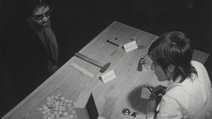 Filmstill aus LES TROIS QUARTS DE LA VIE: Zwei Personen sitzen sich an einem Tisch gegenüber. Auf dem Tisch liegen Kärtchen und diverses Werkzeug.