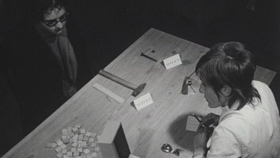 Film still from LES TROIS QUARTS DE LA VIE: Two people are sitting across from each other at a table. There are small cards and various tools on the table.