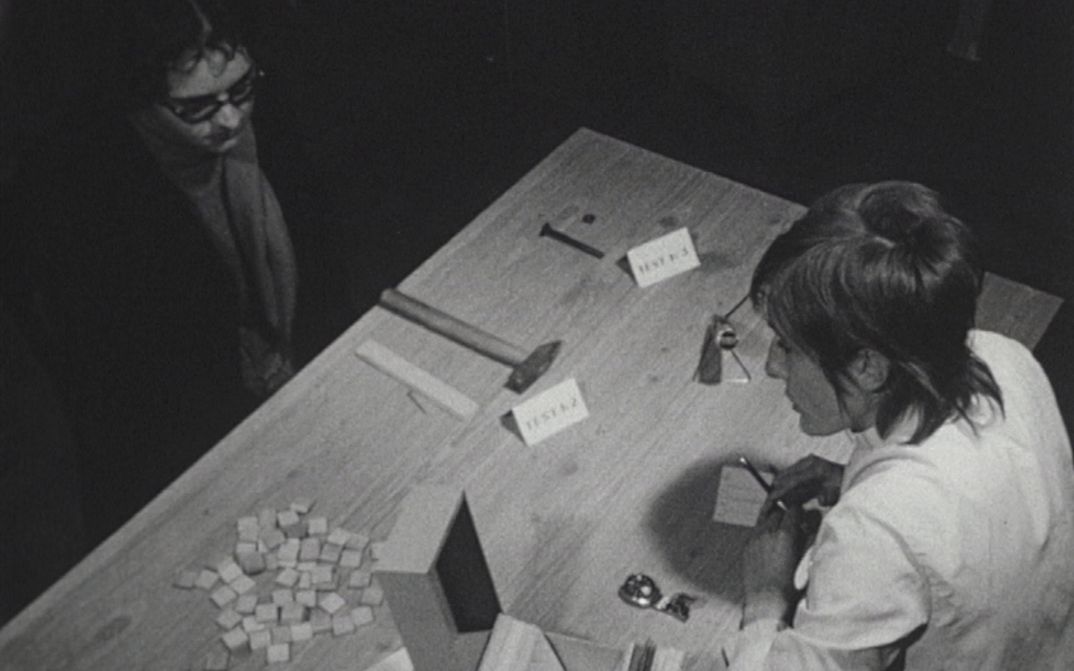 Filmstill aus LES TROIS QUARTS DE LA VIE: Zwei Personen sitzen sich an einem Tisch gegenüber. Auf dem Tisch liegen Kärtchen und diverses Werkzeug.