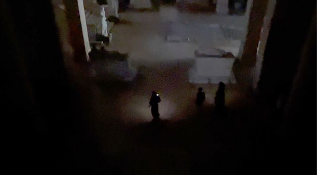 Film still from Joana Hadjithomas & Khalil Joreige’s film “Sarcophagus of Drunken Loves”. People shine some light onto an otherwise blacked-out space, and a statue and staircase can be seen on the left side of the frame. 