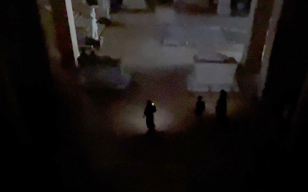 Film still from Joana Hadjithomas & Khalil Joreige’s film “Sarcophagus of Drunken Loves”. People shine some light onto an otherwise blacked-out space, and a statue and staircase can be seen on the left side of the frame. 