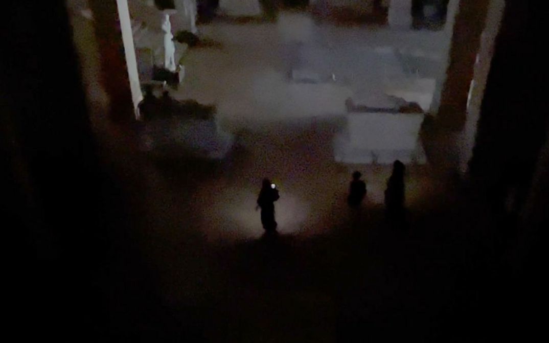 Film still from Joana Hadjithomas & Khalil Joreige’s film “Sarcophagus of Drunken Loves”. People shine some light onto an otherwise blacked-out space, and a statue and staircase can be seen on the left side of the frame.