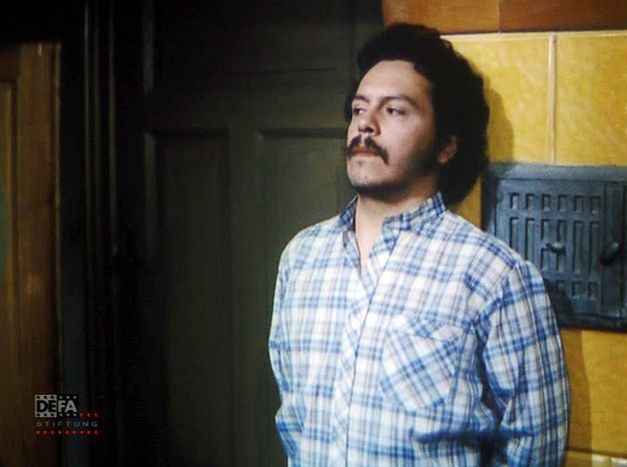 Filmstill aus BLONDER TANGO: Ein dunkelgelockter Mann lehnt an einem Kachelofen.