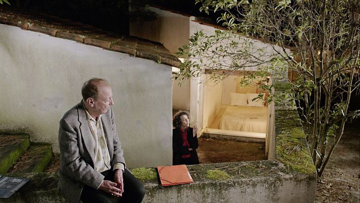 Still from the film "O trio em mi bemol (The Kegelstatt Trio)" by Rita Azevedo Gomes. A man sits on a garden wall. In the background a woman stands smoking at the bottom of the stairs. 
