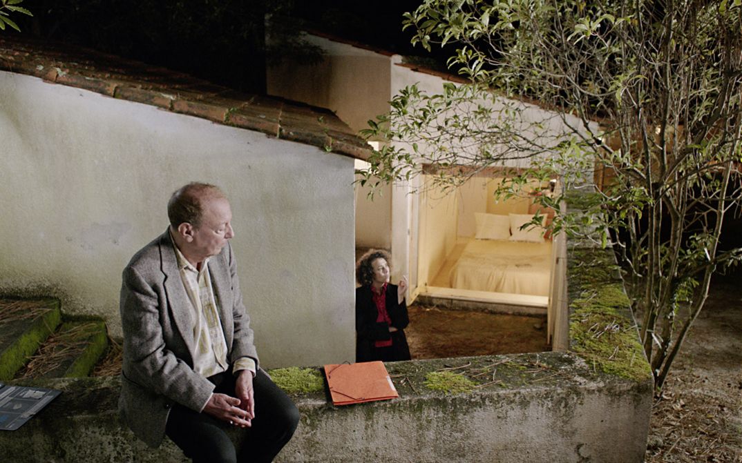 Still from the film "O trio em mi bemol (The Kegelstatt Trio)" by Rita Azevedo Gomes. A man sits on a garden wall. In the background a woman stands smoking at the bottom of the stairs. 