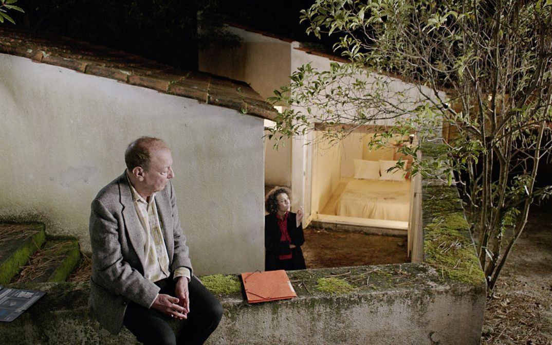 Filmstill aus „O trio em mi bemol (The Kegelstatt Trio)“ von Rita Azevedo Gomes. Ein Mann sitzt auf einer Gartenmauer. Im Hintergrund steht eine Frau am Fuß einer Treppe und raucht.