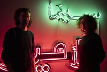 The two are standing in the dark against a wall with neon lights that are shining red and green.