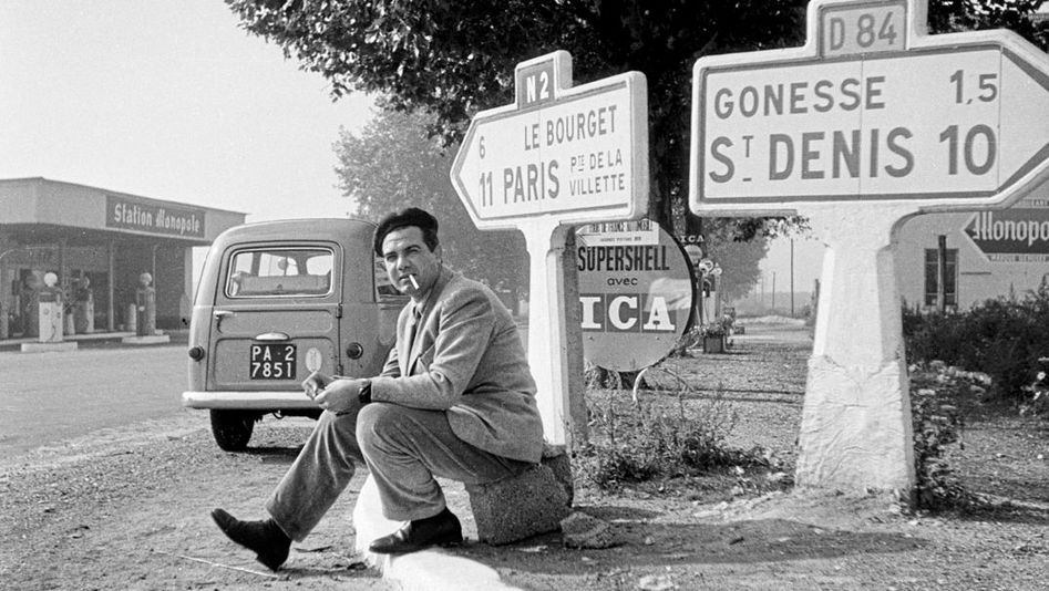 Film still from "Il cassetto segreto" by Costanza Quatriglio. It shows a black and white image of a man with a cigarette in his mouth sitting on the side of the road. Behind him are two road signs pointing the way. In the background is a petrol station.