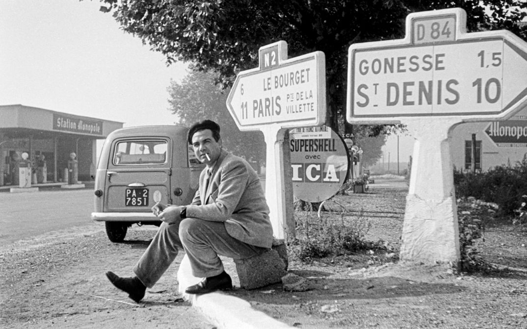 Film still from "Il cassetto segreto" by Costanza Quatriglio. It shows a black and white image of a man with a cigarette in his mouth sitting on the side of the road. Behind him are two road signs pointing the way. In the background is a petrol station.