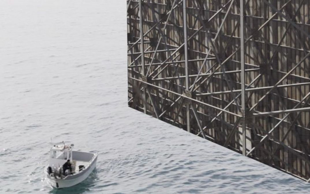Still from the film "Gazing... Unseeing" by Mohamed Abdelkarim. A boat in the sea in front of the back of a billboard.