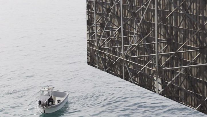 Still from the film "Gazing... Unseeing" by Mohamed Abdelkarim. A boat in the sea in front of the back of a billboard.