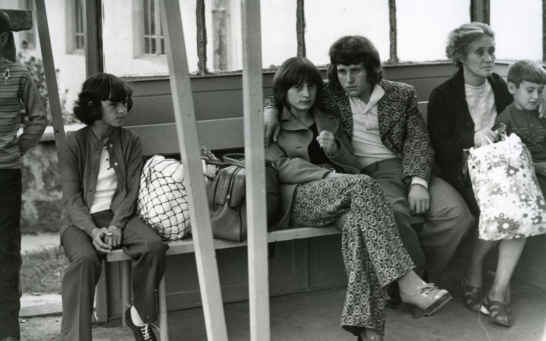 Film still from A COMMONPLACE STORY: Several people are sitting on a bench in a waiting room. In the center is a young couple; she is looking directly at the camera.