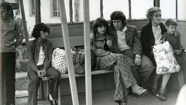 Film still from A COMMONPLACE STORY: Several people are sitting on a bench in a waiting room. In the center is a young couple; she is looking directly at the camera. 