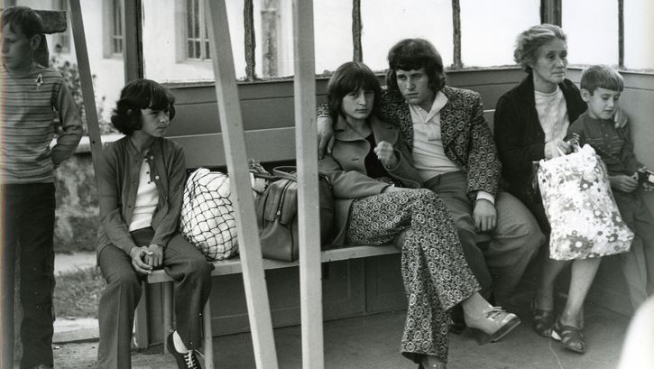 Film still from A COMMONPLACE STORY: Several people are sitting on a bench in a waiting room. In the center is a young couple; she is looking directly at the camera. 