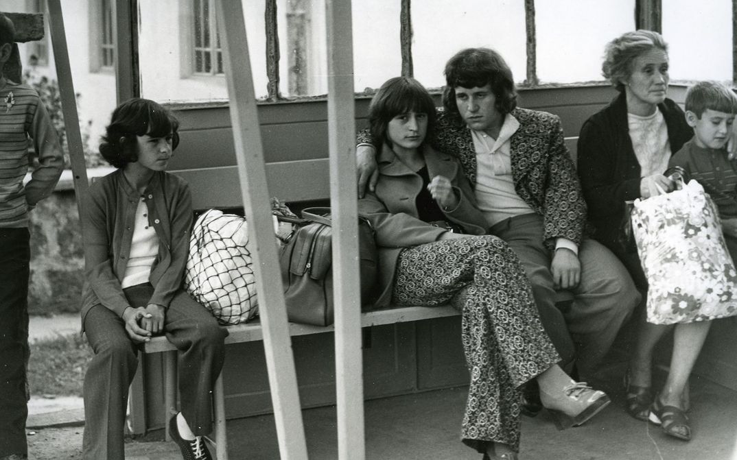 Film still from A COMMONPLACE STORY: Several people are sitting on a bench in a waiting room. In the center is a young couple; she is looking directly at the camera. 