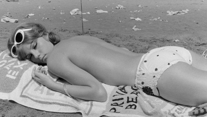 Film still from IO LA CONOSCEVO BENE: A young woman, dressed only in bikini bottoms, lies on her stomach on a bath towel on the beach.