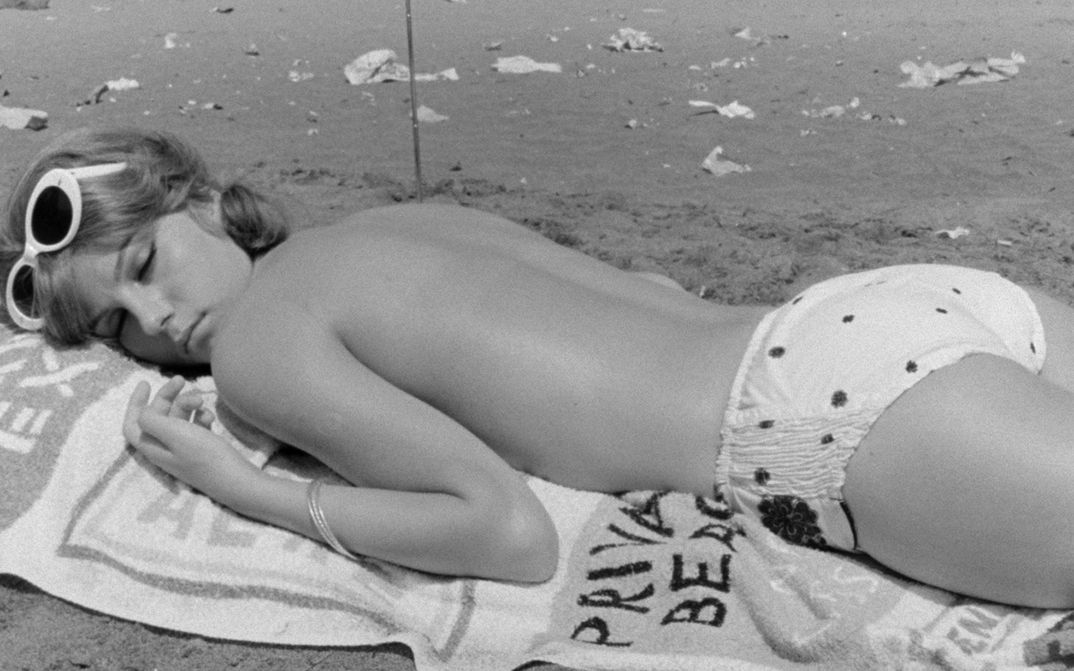 Film still from IO LA CONOSCEVO BENE: A young woman, dressed only in bikini bottoms, lies on her stomach on a bath towel on the beach.