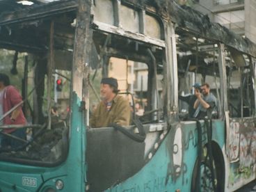 35mm colour photo of a bus that has been burned and vandalized. A man sits in the driver’s seat, smiling, while others inside the bus take photos with their cameras.