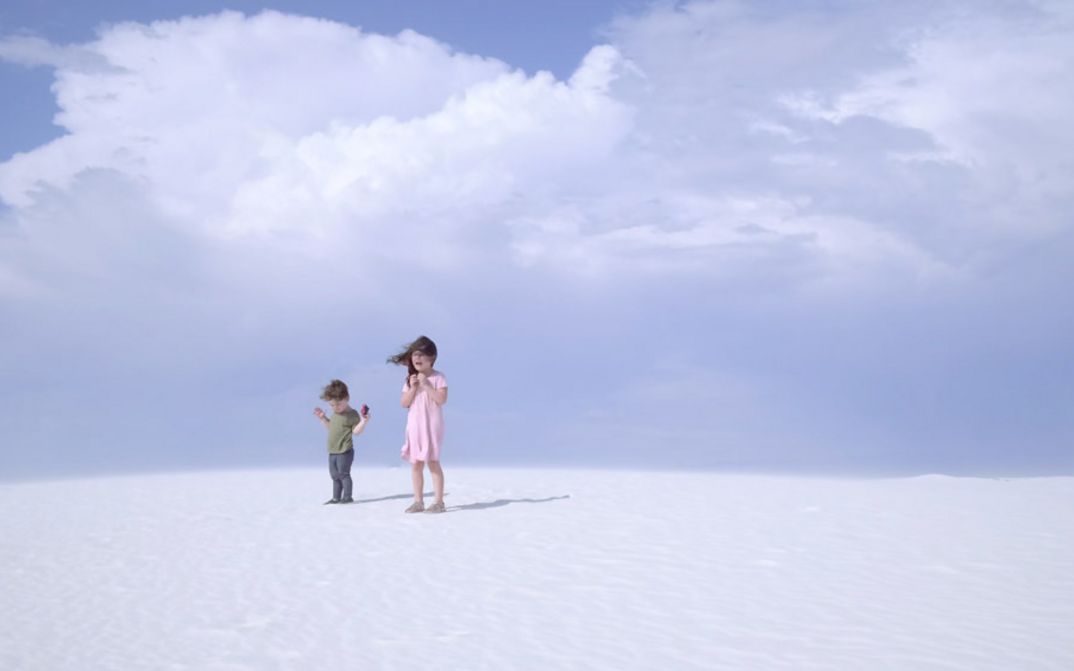Filmstill aus „Nuclear Family“ von Erin Wilkerson und Travis Wilkerson. Zwei Kinder stehen auf einer Sanddüne vor blauem Himmel.