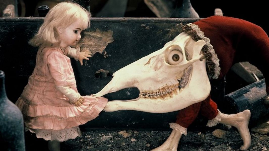 Filmstill aus ALICE: Eine Puppe mit blondem Haar und rosafarbenen Kleid, neben ihr eine groteske Figur, mit einem Tierschädel, der das Kleid in den Mund nimmt.